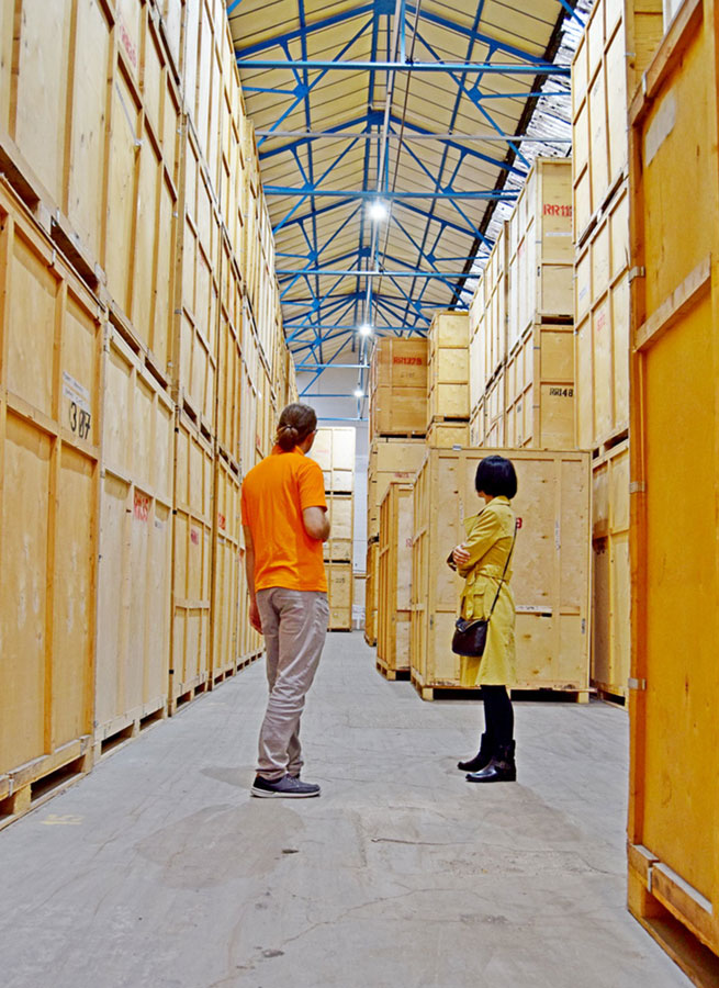 A large storage warehouse filled with wooden crates, with a worker and a woman talking.