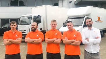 Five men in orange "Removals & StorageX" shirts pose with arms crossed in front of removal trucks.