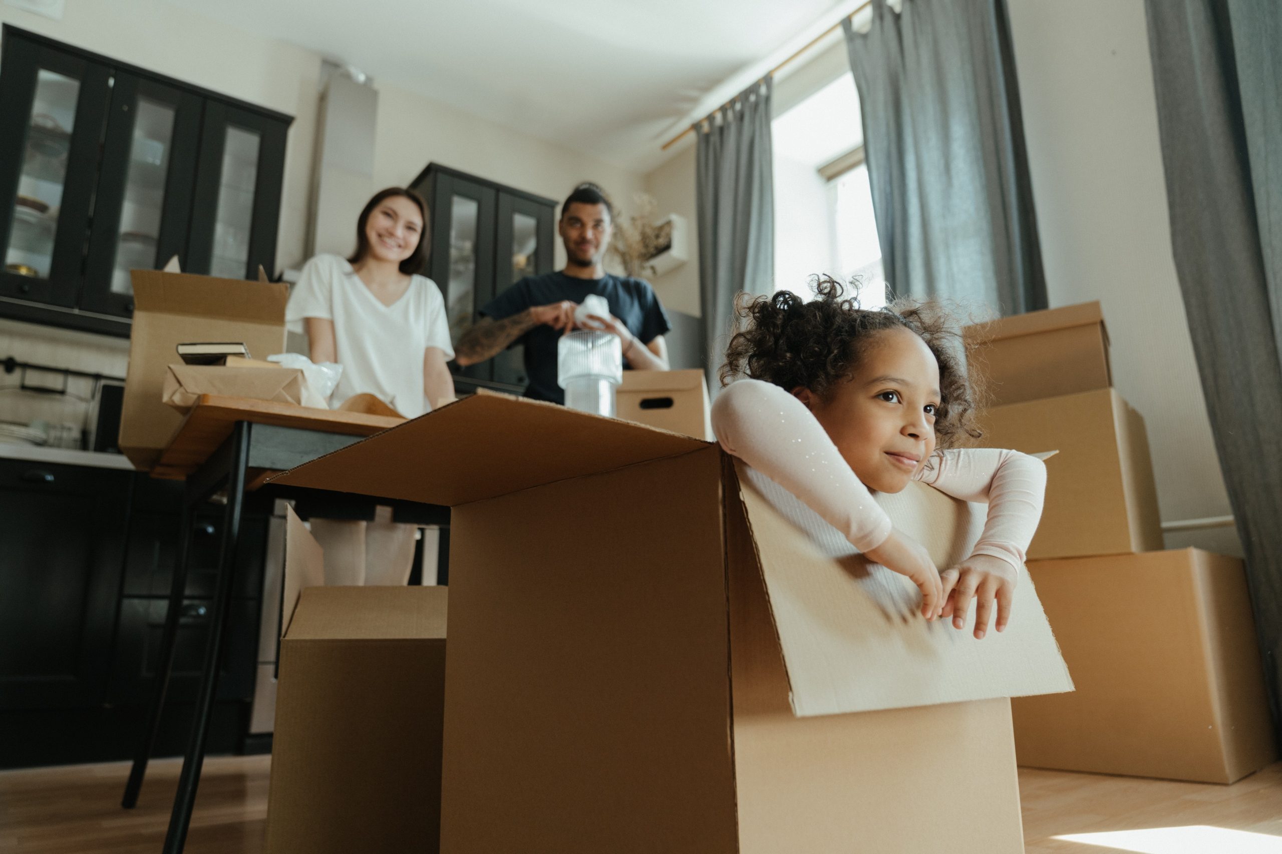 A child playing inside a cardboard box, while two adults pack in the background.