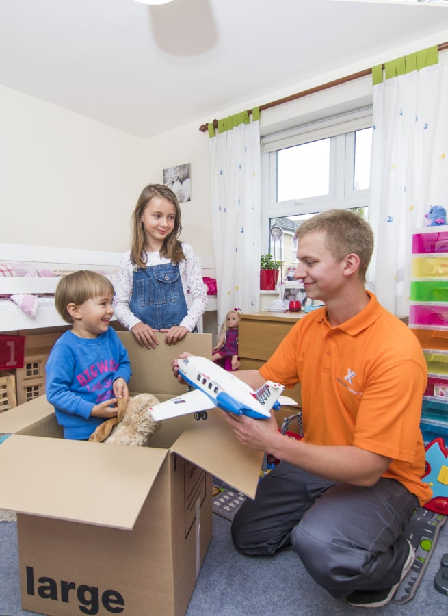 A "Removals and StorageX" worker unpacking a red book from a box in a living room.