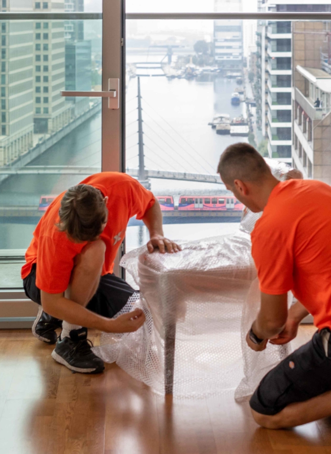 Two workers wrap a chair in bubble wrap near large windows with a city view.